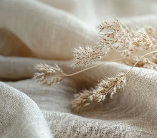 A close-up, minimalist photo of delicate dried pampas grass stems with feathery tops resting on soft, wrinkled light beige linen fabric.