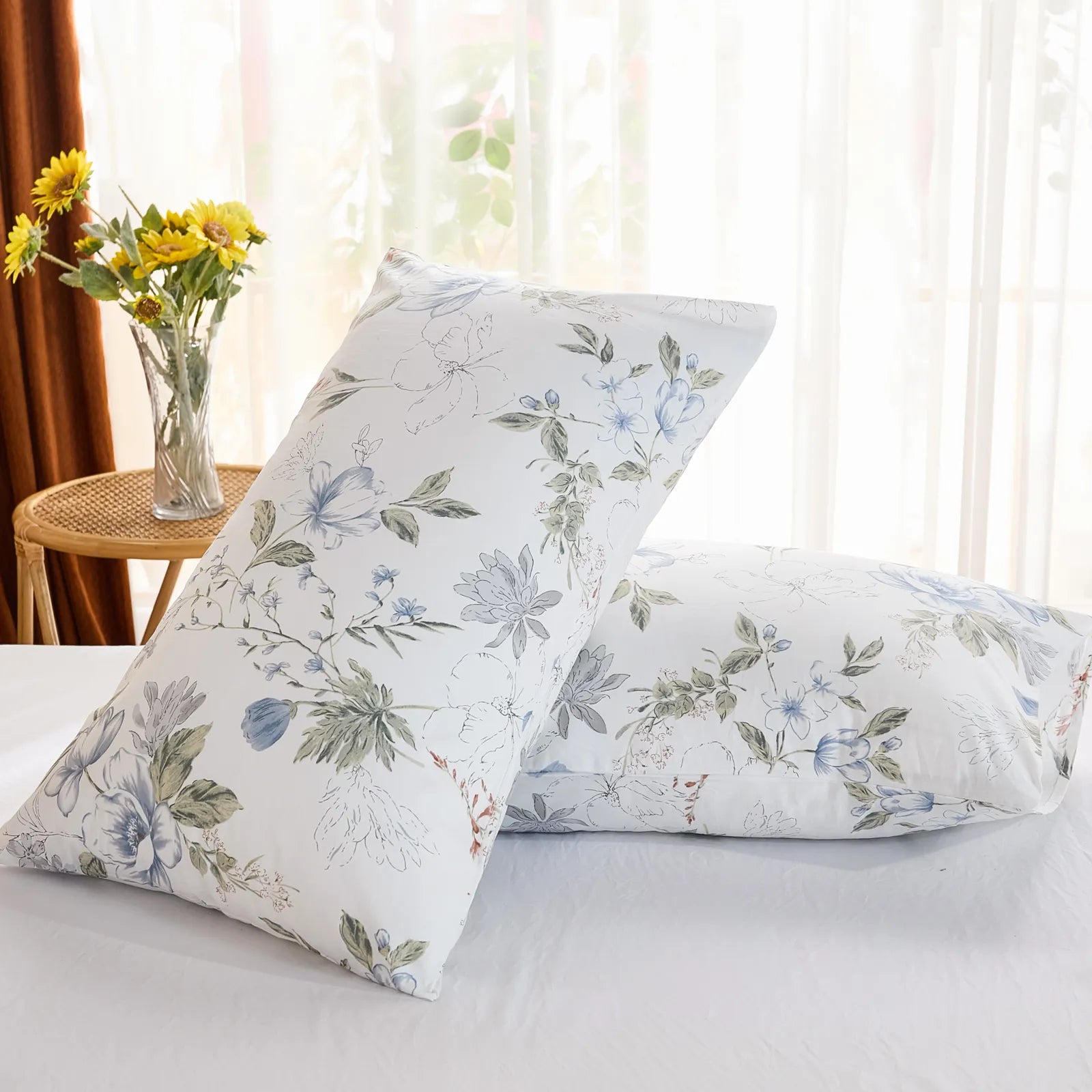 Two cotton pillowcases with a light blue botanical print, shown in bright natural light next to a window.
