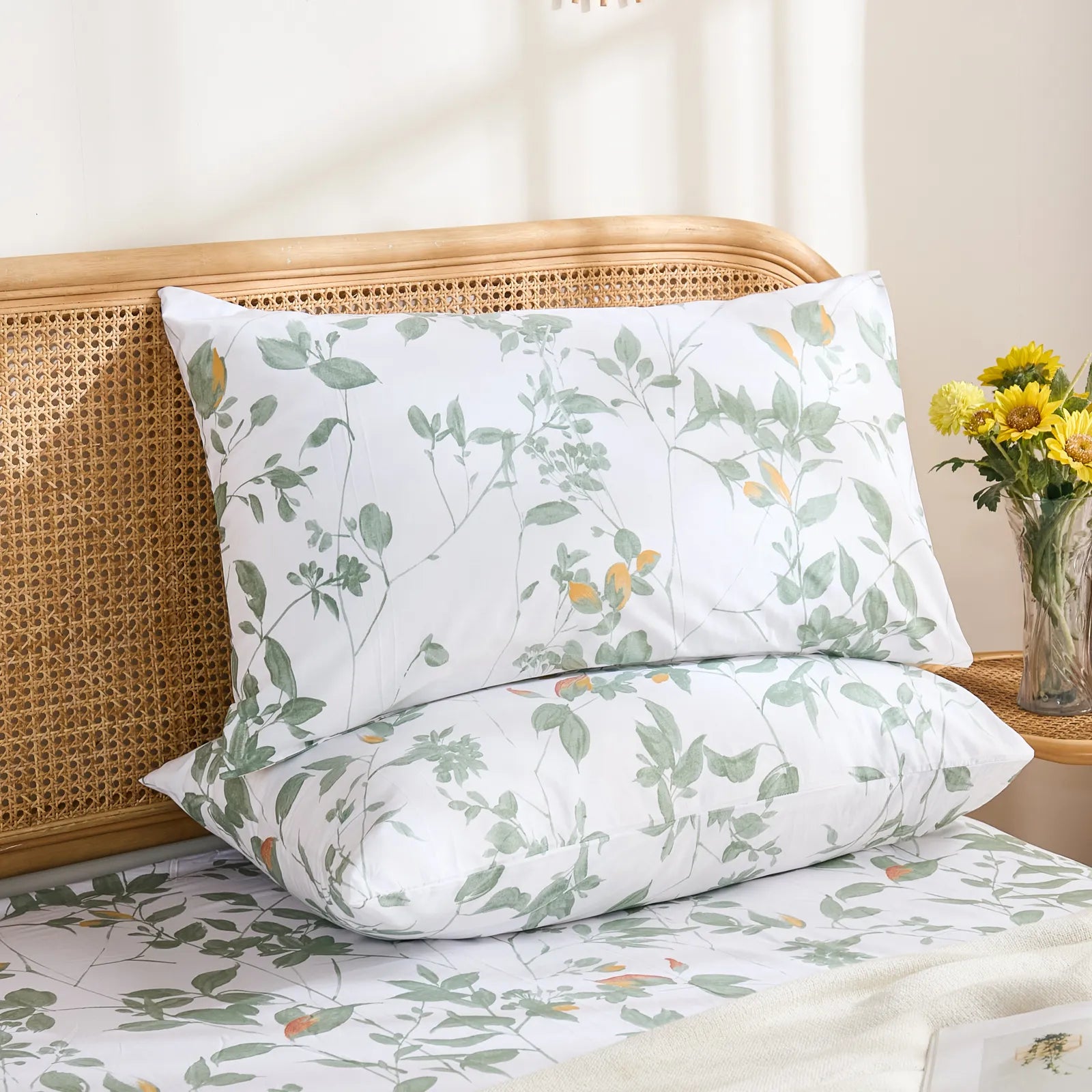 Two cotton pillowcases featuring a serene green leafy print, stacked on a matching fitted sheet in a bright bedroom setting.