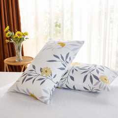 An angled view of two white cotton pillowcases with a yellow and grey floral print, propped up against each other on a bed near a window