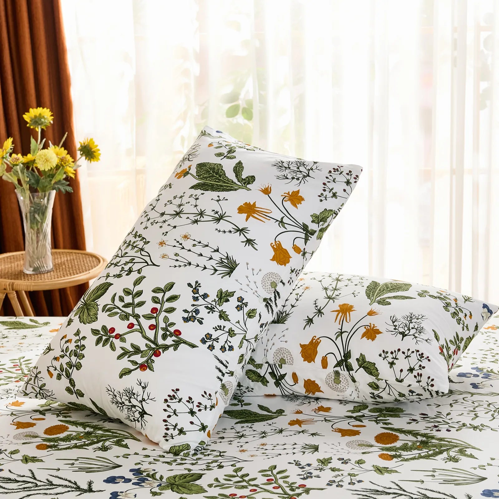 An angled shot of two white cotton botanical pillowcases leaning against each other on a bed, with a vase of yellow flowers in the background.