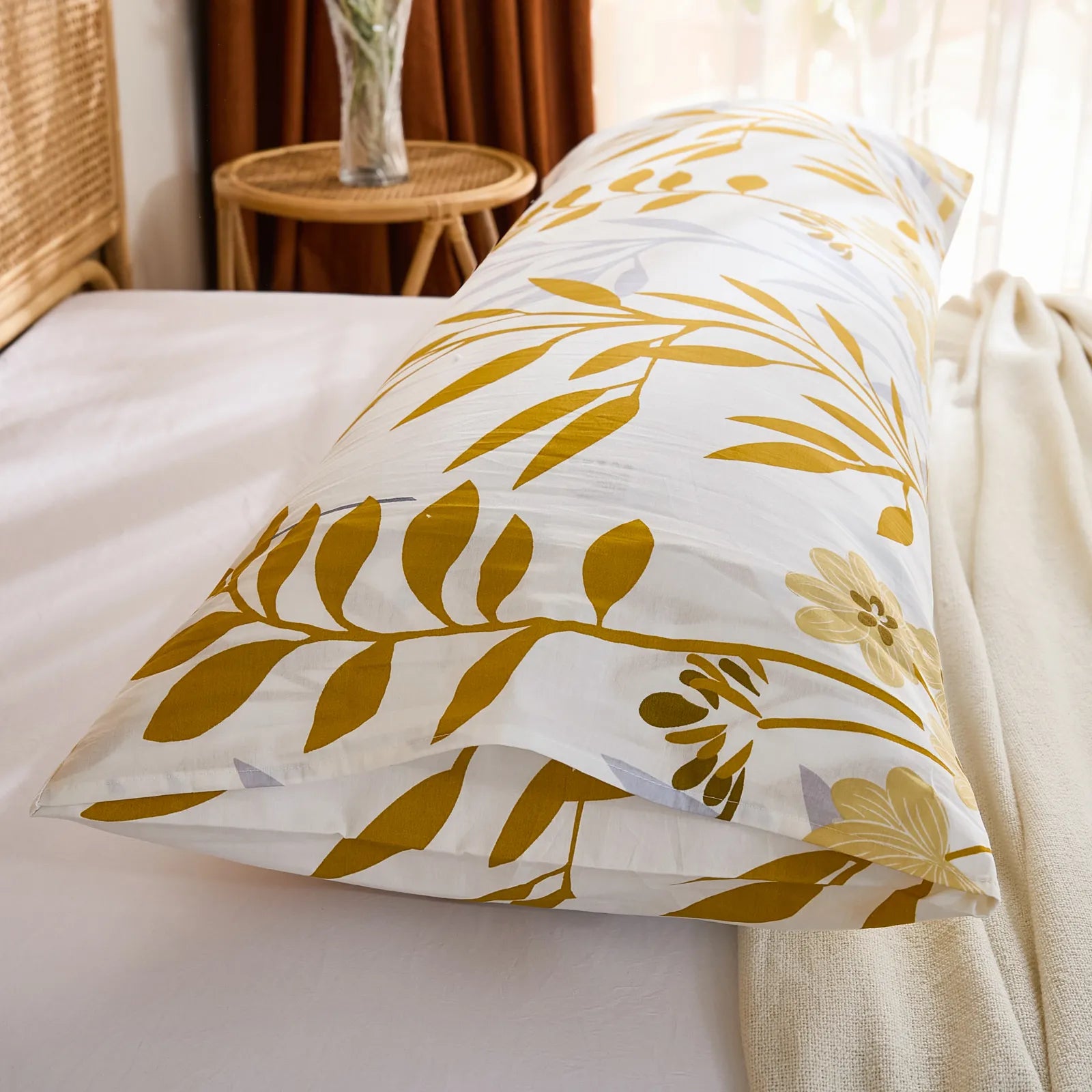 An angled view of a long white cotton body pillow, highlighting the vibrant yellow leaf print against the soft natural lighting from a window.