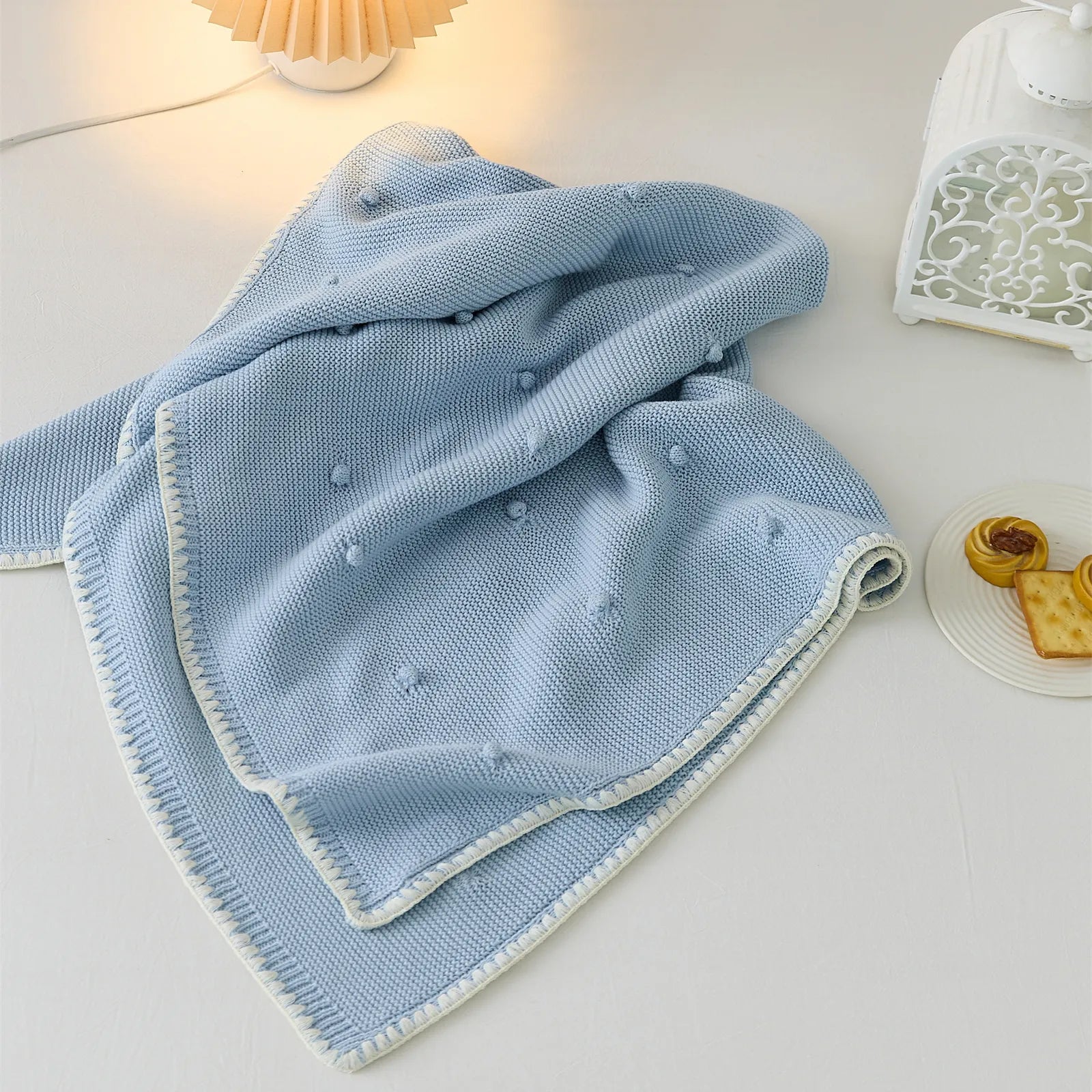 Baby blue cotton bobble throw draped on white bed next to small white cake and crackers