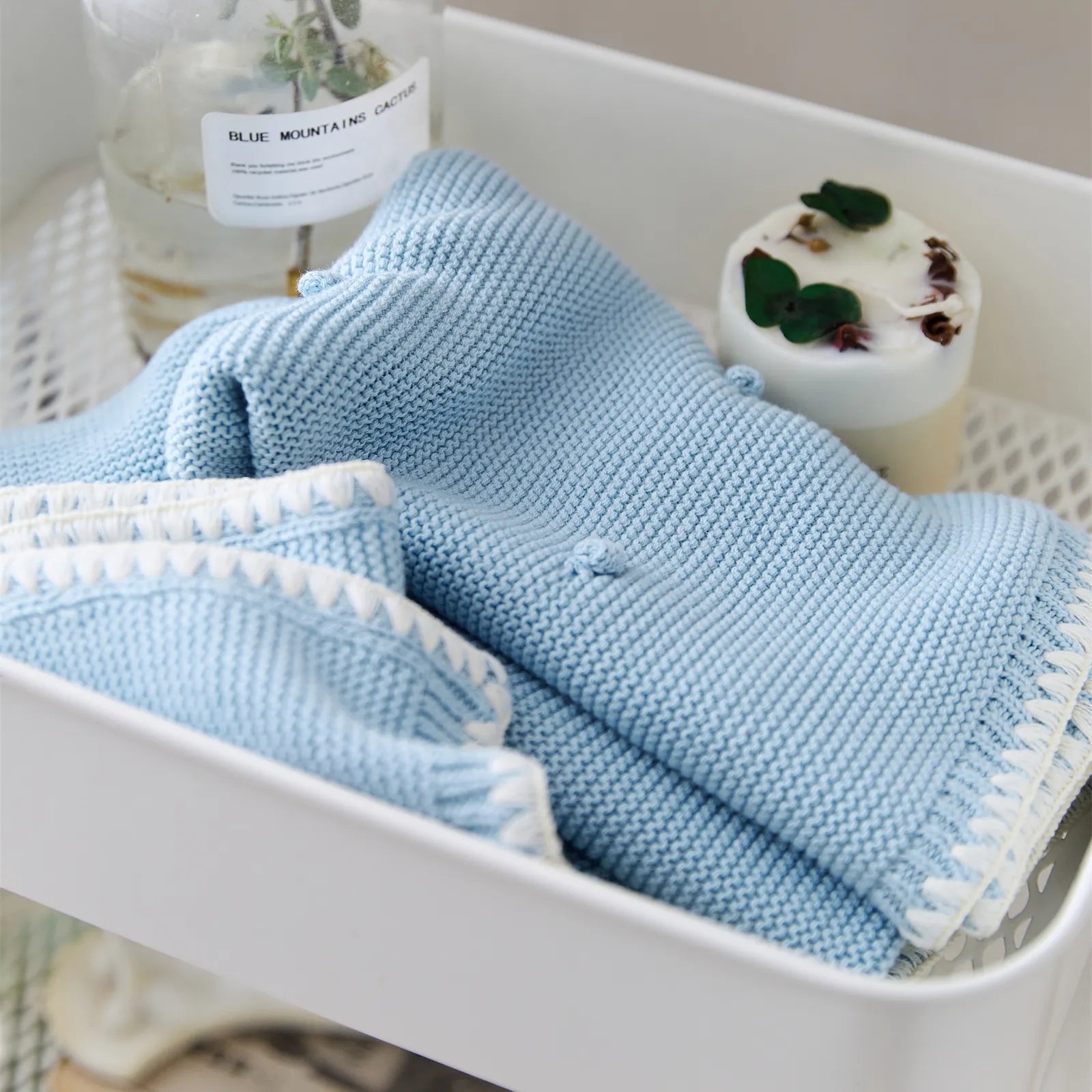 Folded baby blue cotton knit throw placed in a white storage cart next to a glass vase and candle
