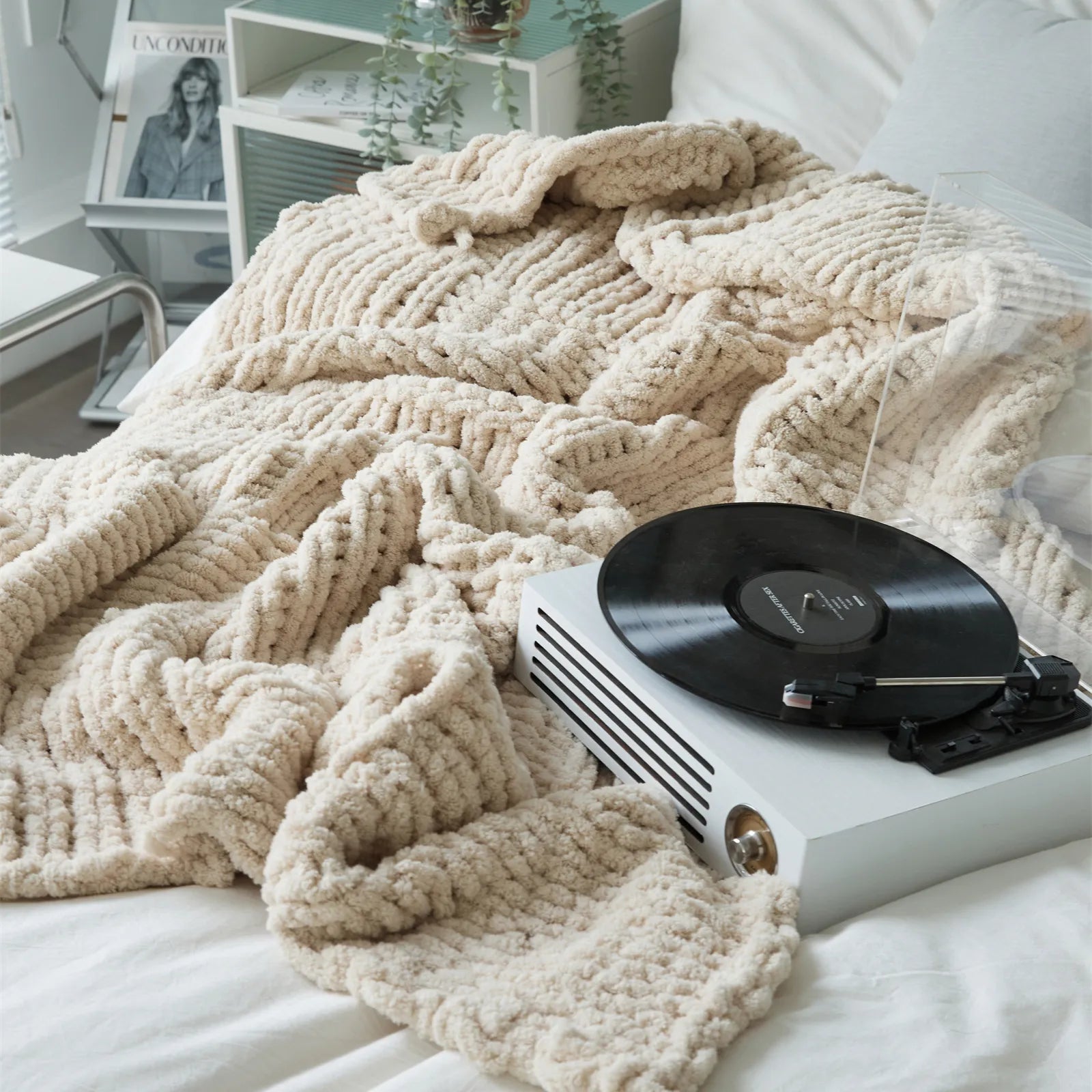 A cozy beige chunky knit blanket styled on a bed next to a record player, creating a relaxed, modern vibe.
