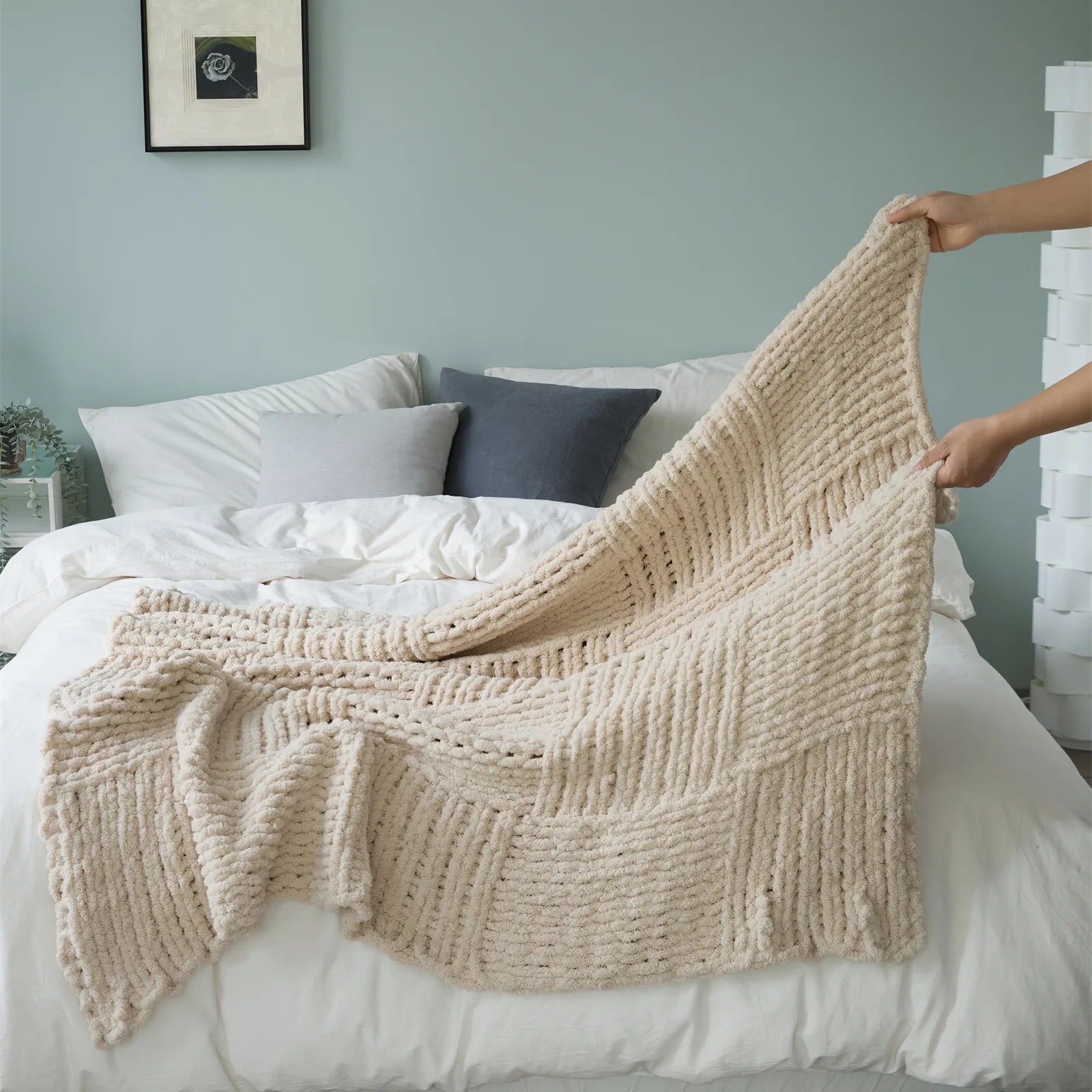 A pair of hands holding up a soft, beige chunky knit throw blanket, highlighting its cozy texture and style on a bed