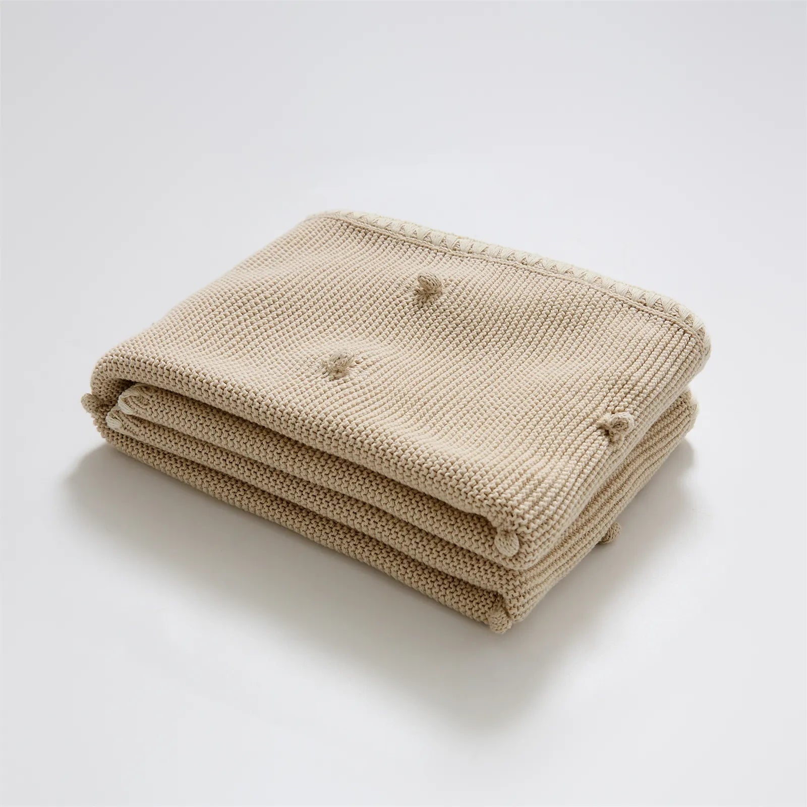 Neatly folded beige cotton knit throw blanket with cream fringe trim and bobble texture