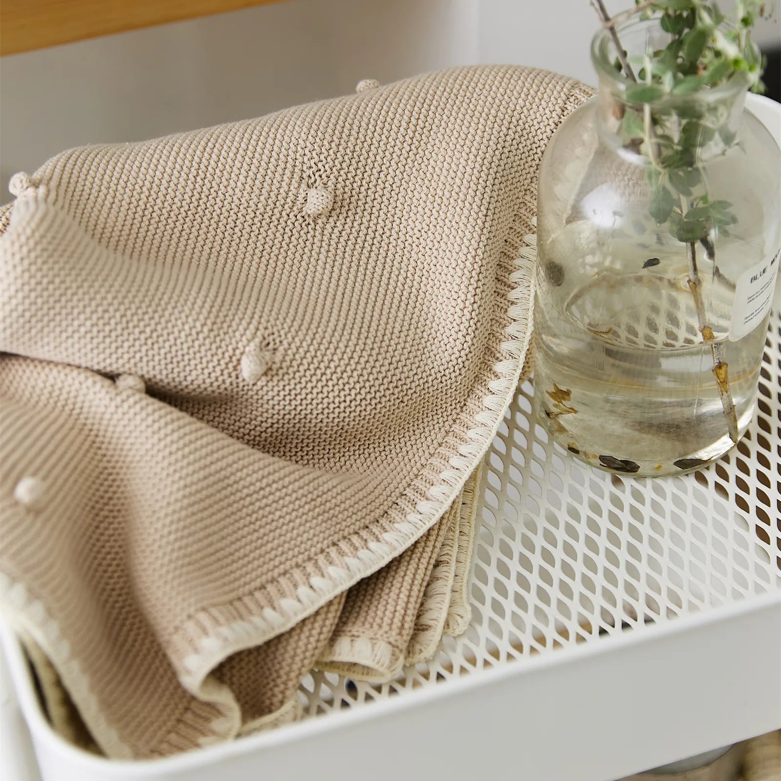 Beige cotton bobble throw draped over a white cart next to a glass vase with greenery