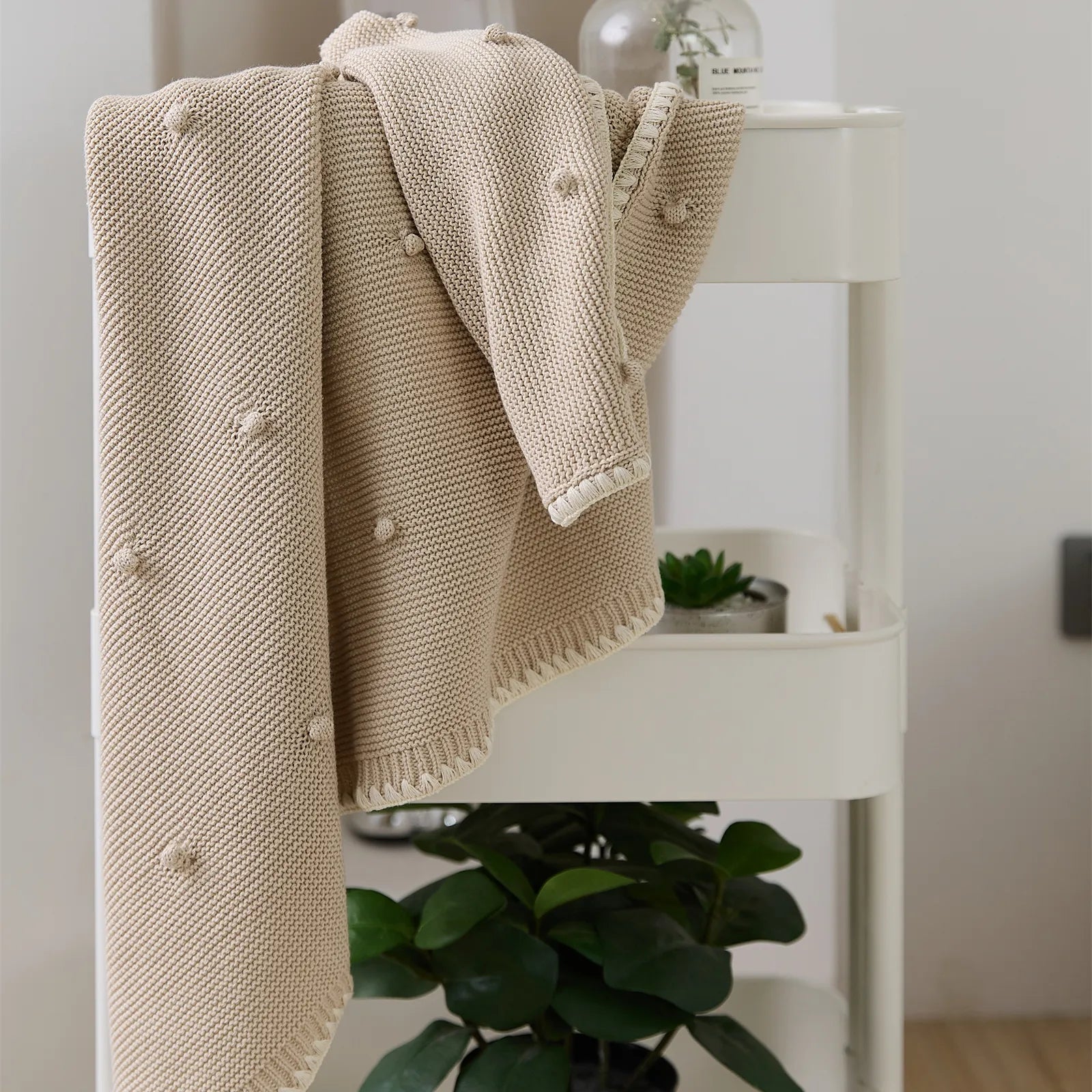 Beige cotton bobble throw blanket hanging on white storage cart with small plants