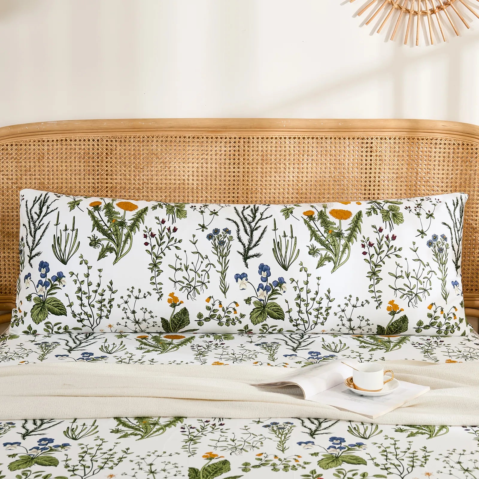 A single long cotton body pillow with a botanical print, placed on a bed with matching sheets and a rattan headboard.