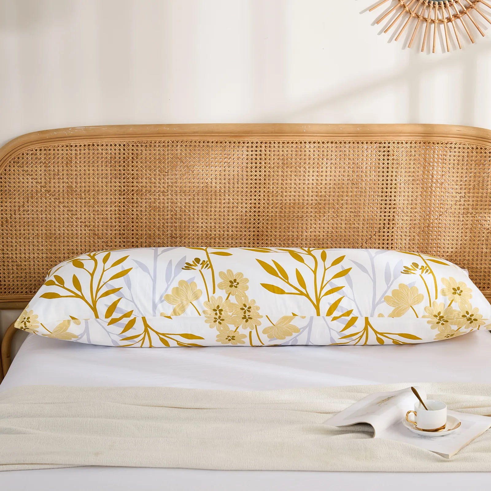 A single long cotton body pillow with a striking yellow leaf and subtle floral botanical print, placed on a bed with a rattan headboard.
