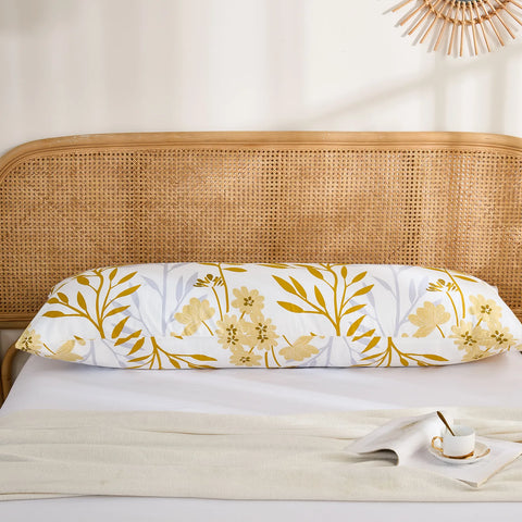 A single long cotton body pillow with a striking yellow leaf and subtle floral botanical print, placed on a bed with a rattan headboard.