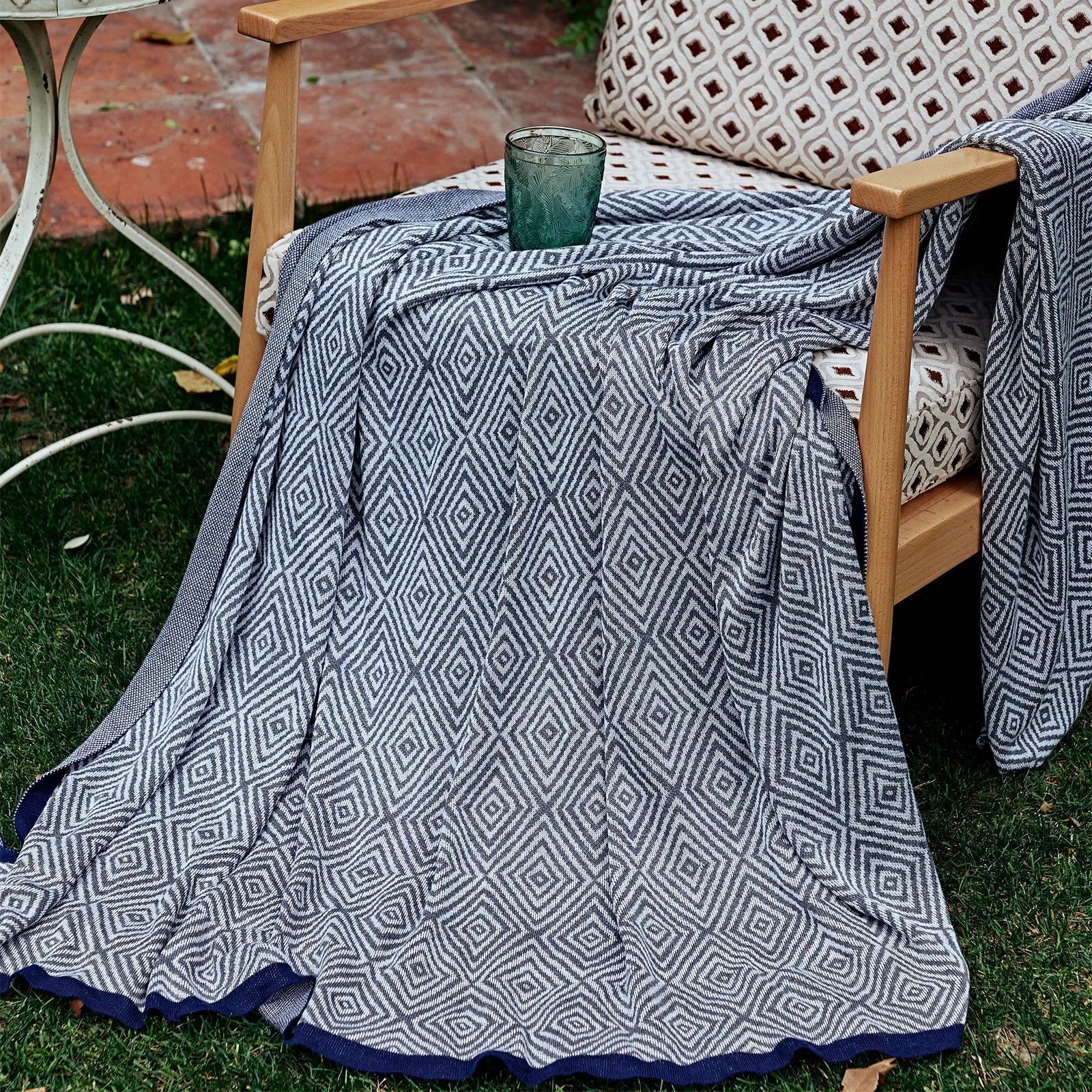 Charcoal-grey diamond pattern throw with a navy border draped over a wooden chair in an outdoor garden setting.