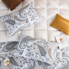 A wide shot of a pair of cotton grey paisley pillowcases on a bed, with a light-colored throw blanket and a decorative rattan headboard. 