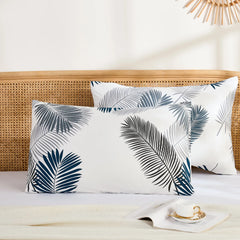 A wide view of two cotton pillowcases with a tropical leaf pattern, placed on a bed with a wicker headboard, a book, and a teacup.