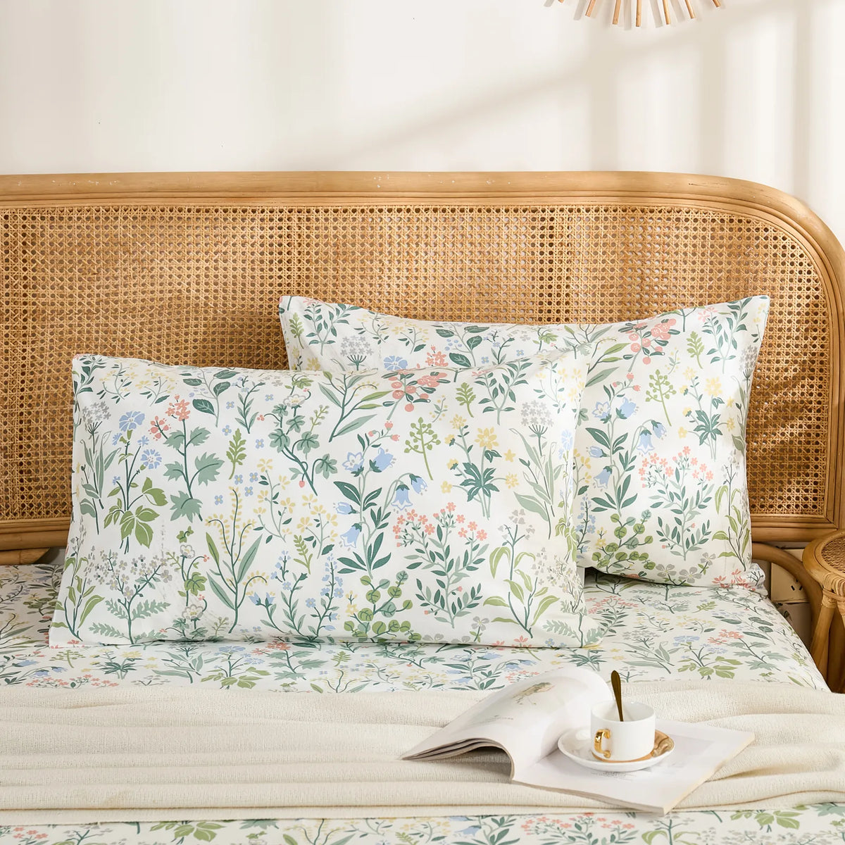 Two cotton meadow flower pillowcases are neatly arranged on a bed with a rattan headboard, styled with a book and a teacup.