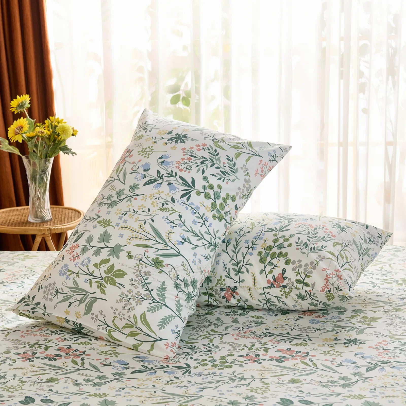 A pair of cotton meadow print pillowcases are propped on a bed, with a sunlit window and yellow sunflowers providing a warm, natural backdrop.