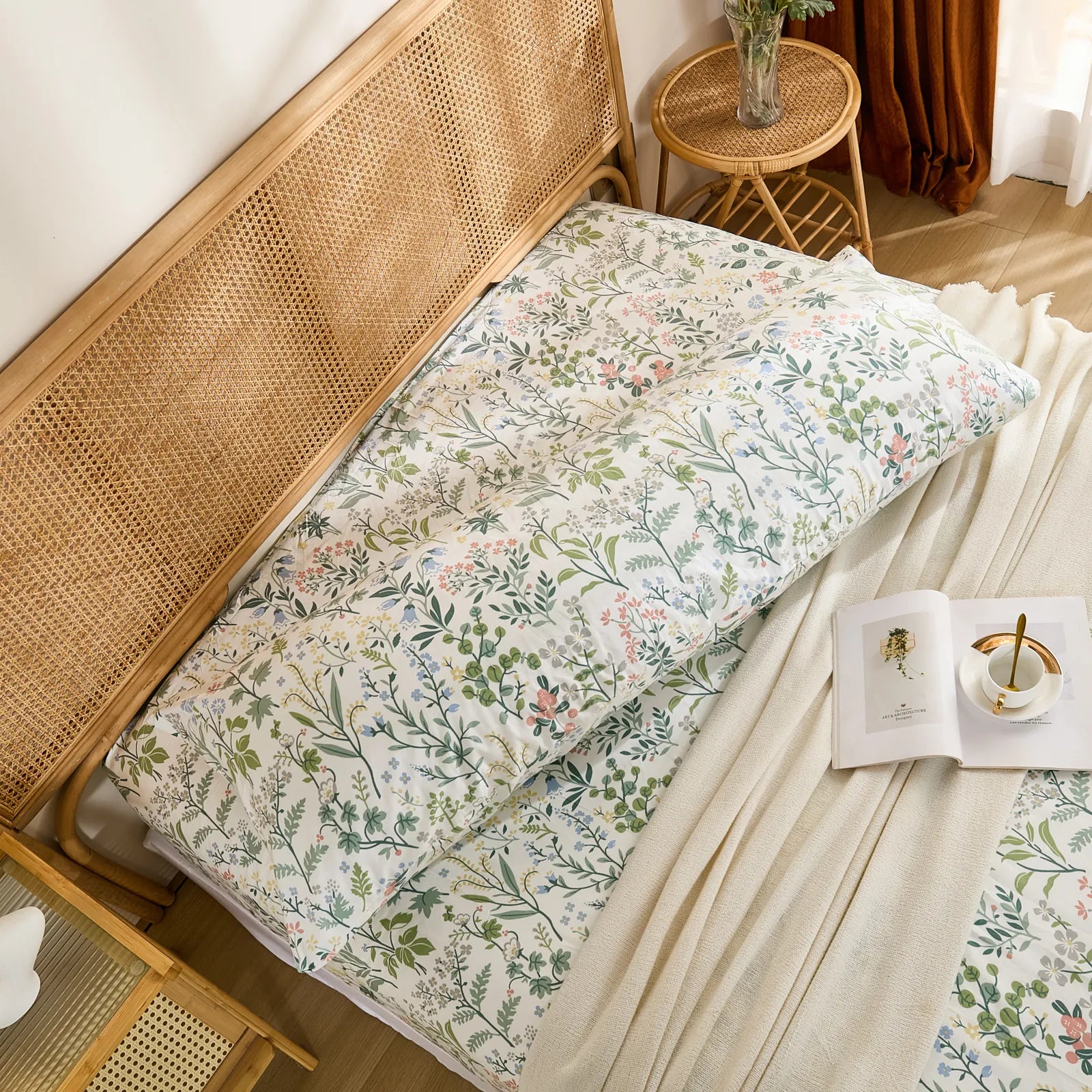 An overhead view of a cotton wildflower body pillowcase on a bed, styled with a book and a teacup.