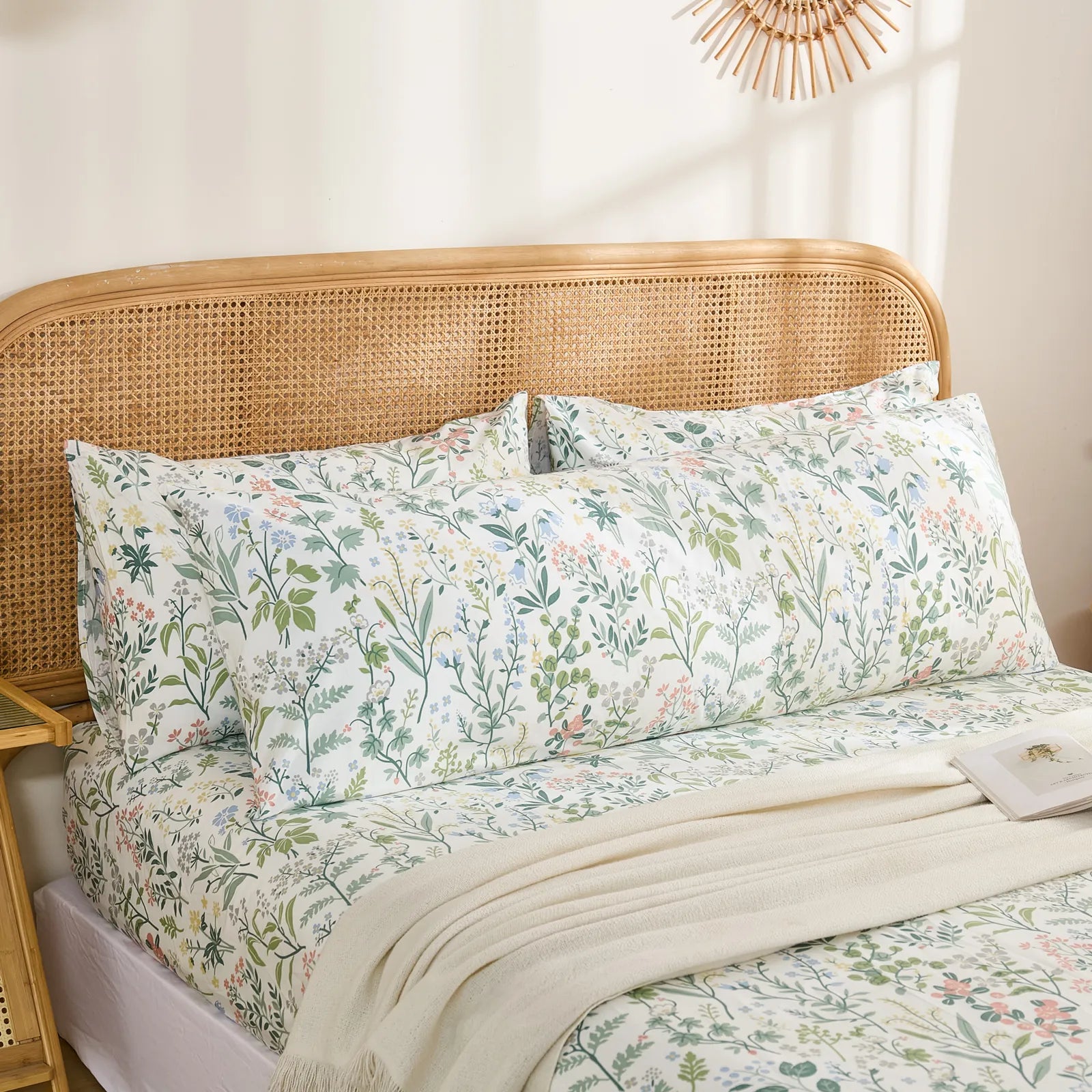 A long cotton body pillowcase with a wildflower print is styled on a bed, with a white throw blanket and a rattan headboard.
