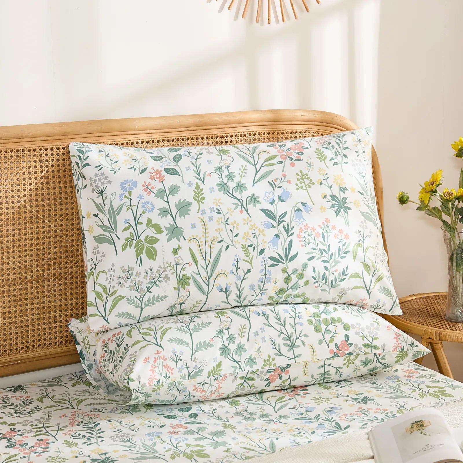 A pair of cotton wildflower print pillowcases are stacked on a bed, showcasing the delicate floral pattern against a light-colored throw blanket and a rattan headboard.