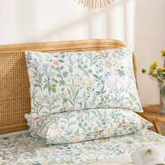 A pair of cotton wildflower print pillowcases are stacked on a bed, showcasing the delicate floral pattern against a light-colored throw blanket and a rattan headboard.