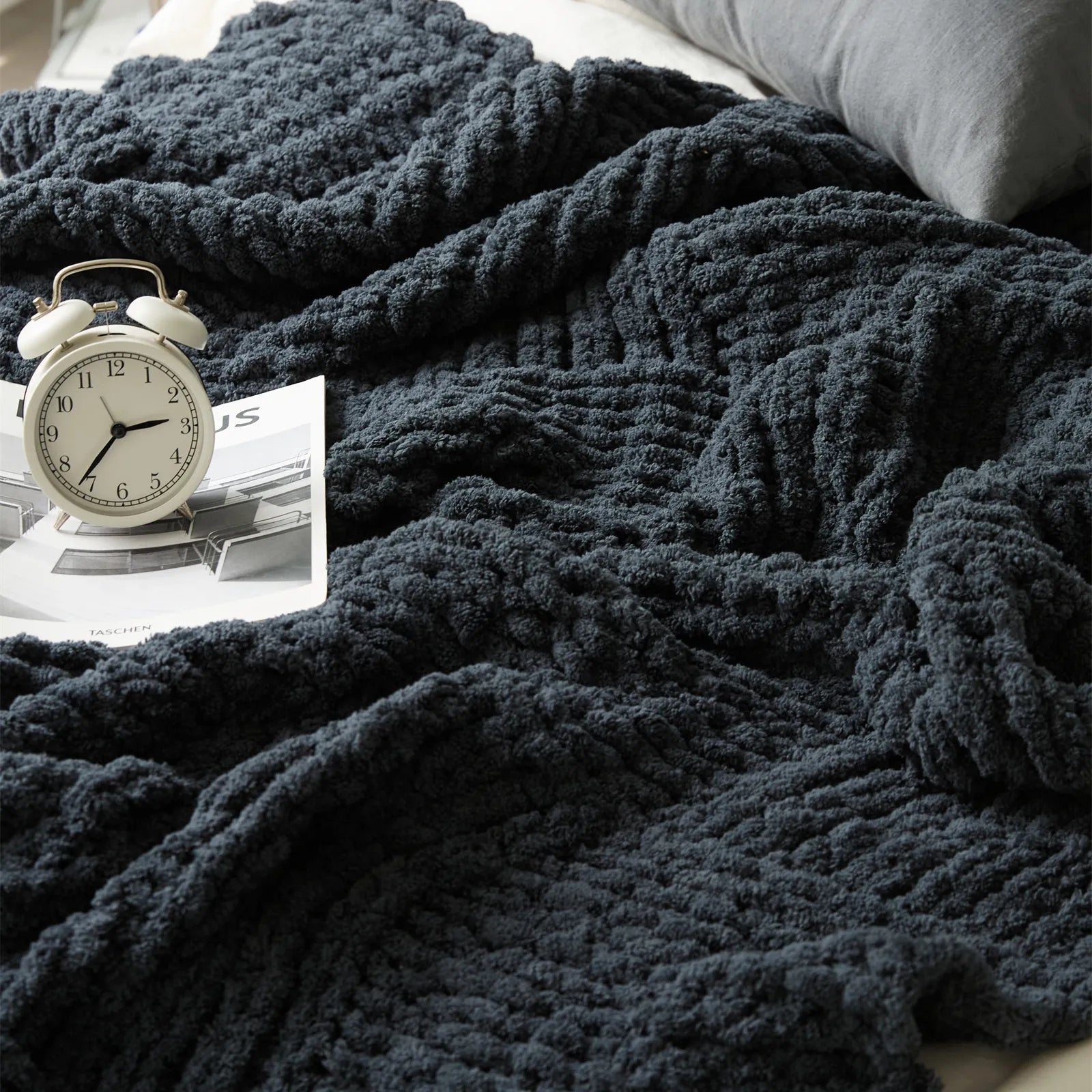 Hands are arranging a plush dark gray chunky knit blanket on a white bed, emphasizing its textured pattern and rich color.