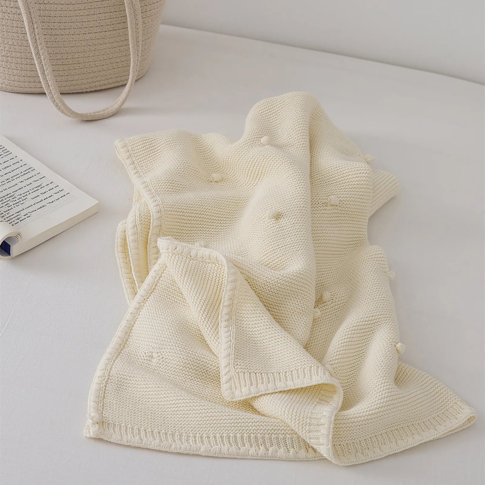 Cream white cotton knit throw blanket loosely folded beside an open book on a bed