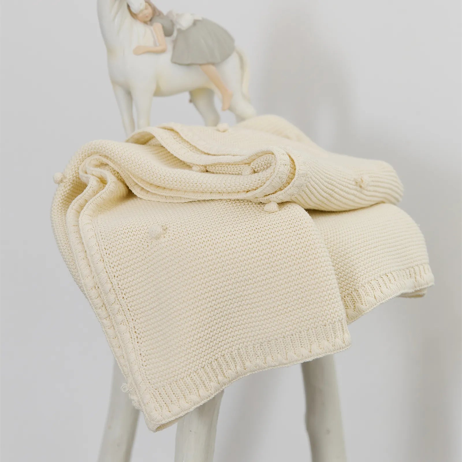 Cream white cotton bobble knit throw draped on a white stool next to a white ceramic figurine