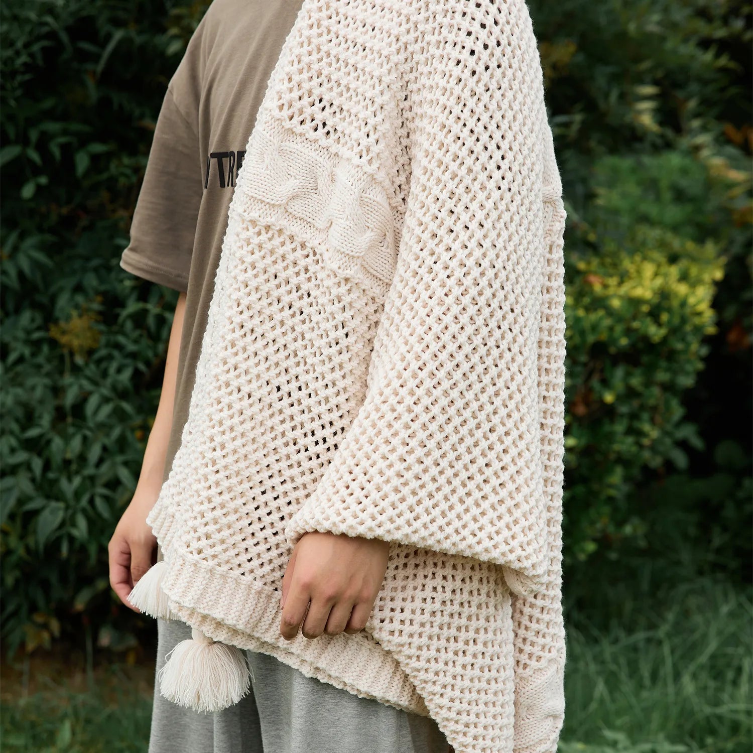 Person wrapped in a cream-white knit throw blanket with tassels, highlighting the open-knit texture outdoors.