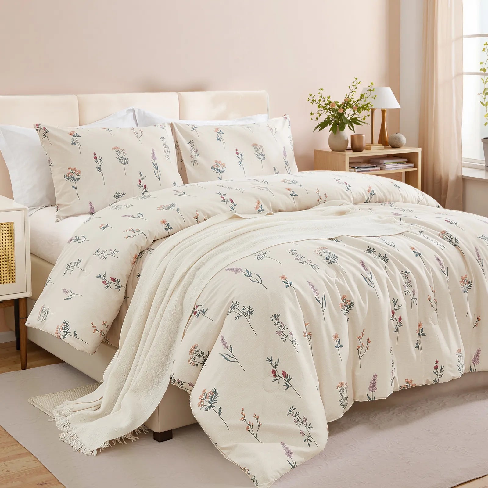 Angled view of a bed with a cream-colored floral comforter and a beige blanket, with a white and rattan nightstand