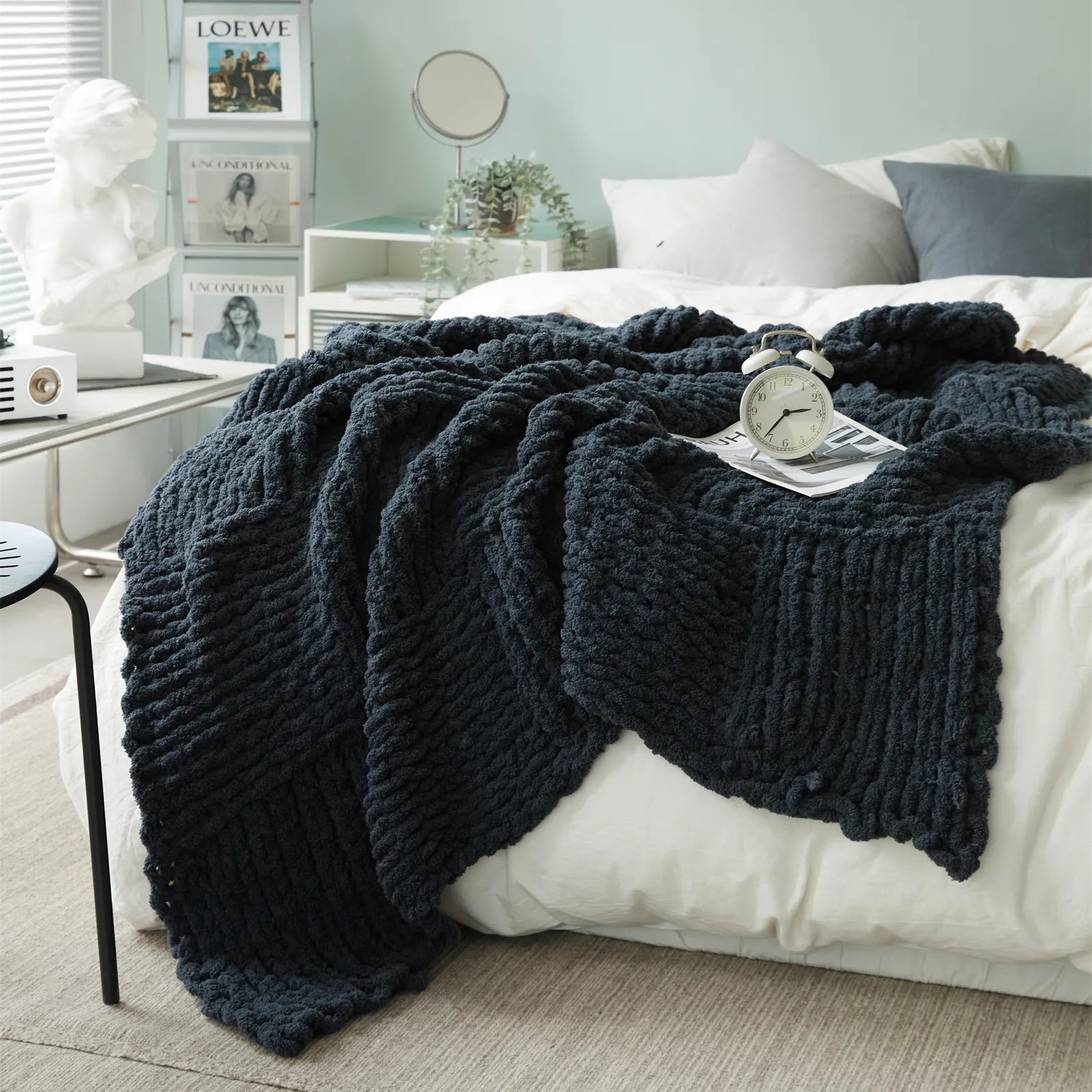 A dark gray chunky knit blanket is spread out on a white bed, adding a deep, calming color and soft texture to the room.