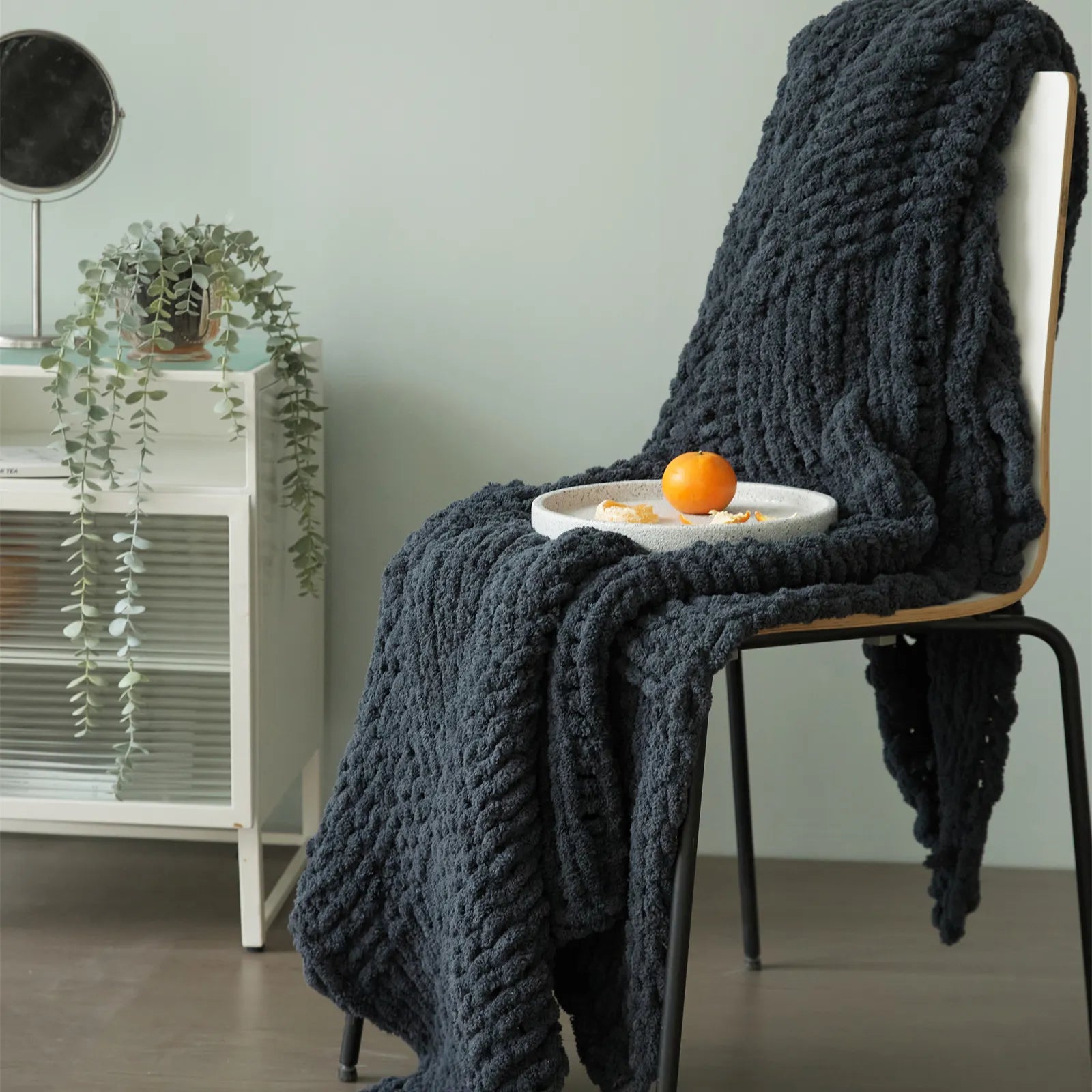A thick dark gray chunky knit blanket draped over a chair, with a plate of fruit on top, showcasing its cozy, stylish appeal.