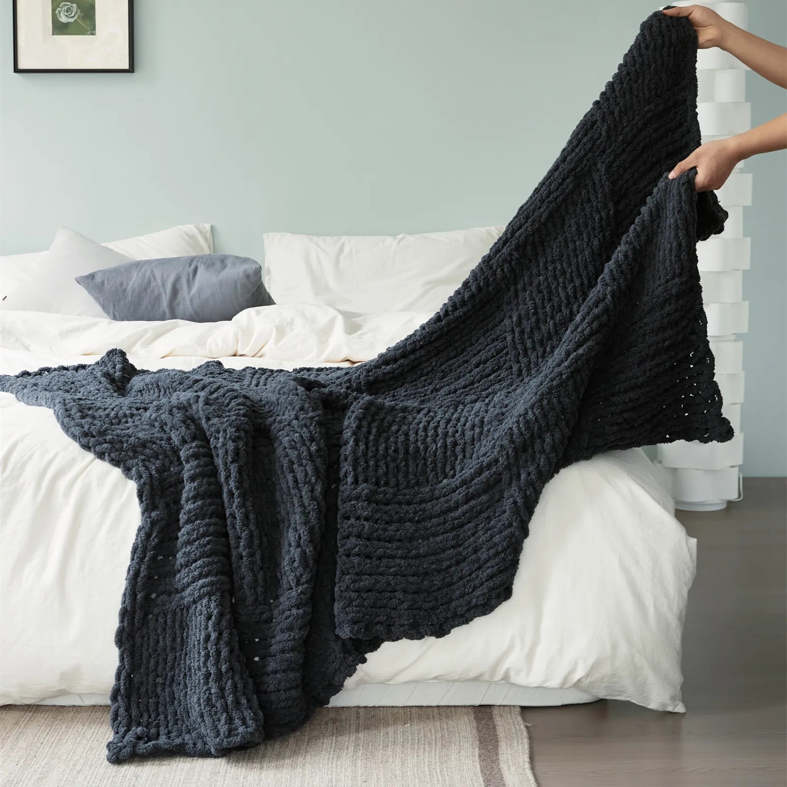 A cozy dark gray chunky knit blanket styled on a bed with an alarm clock and magazine for a comfortable, relaxing atmosphere.