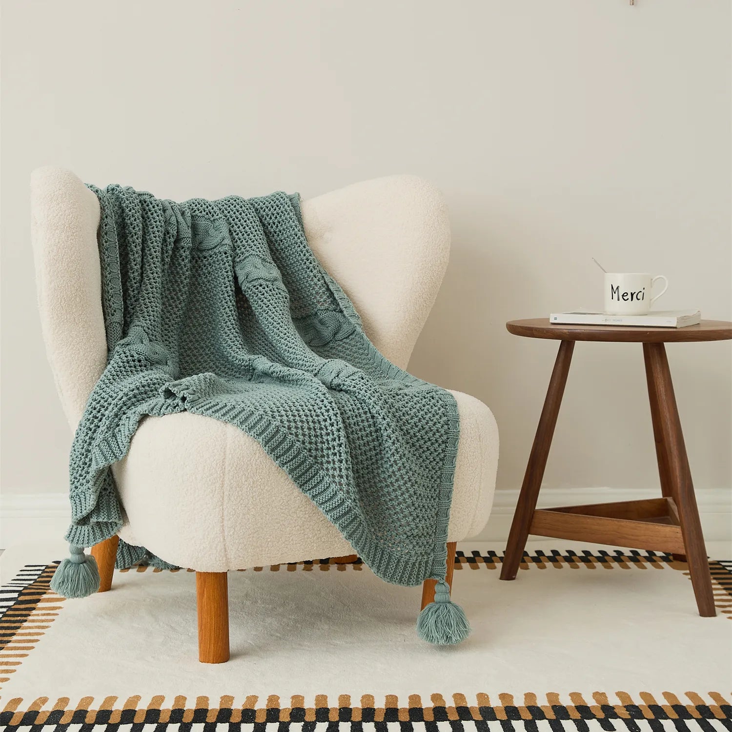 Sage-green knit throw blanket with tassels draped over a cream teddy armchair next to a wooden side table.