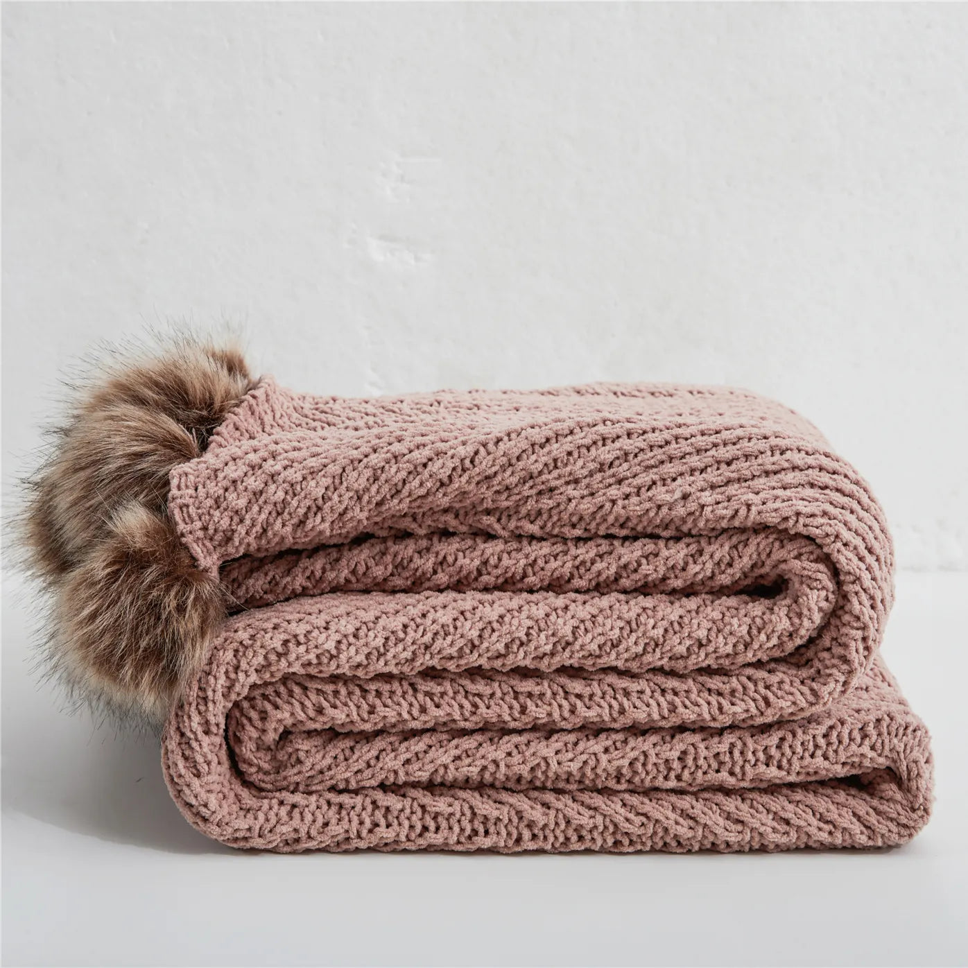 Folded dusty pink chunky knit throw blanket with brown faux fur pom-poms