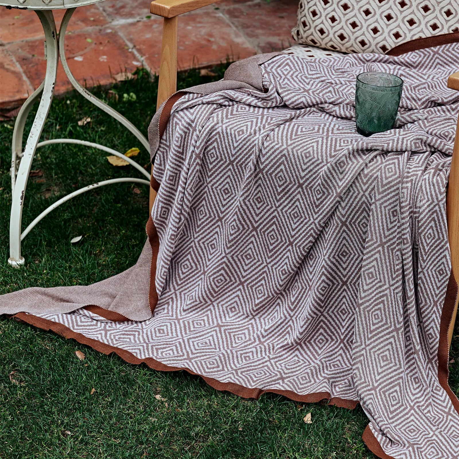 Dusty-pink diamond pattern throw draped over a wooden chair outdoors on the grass, next to a small side table.