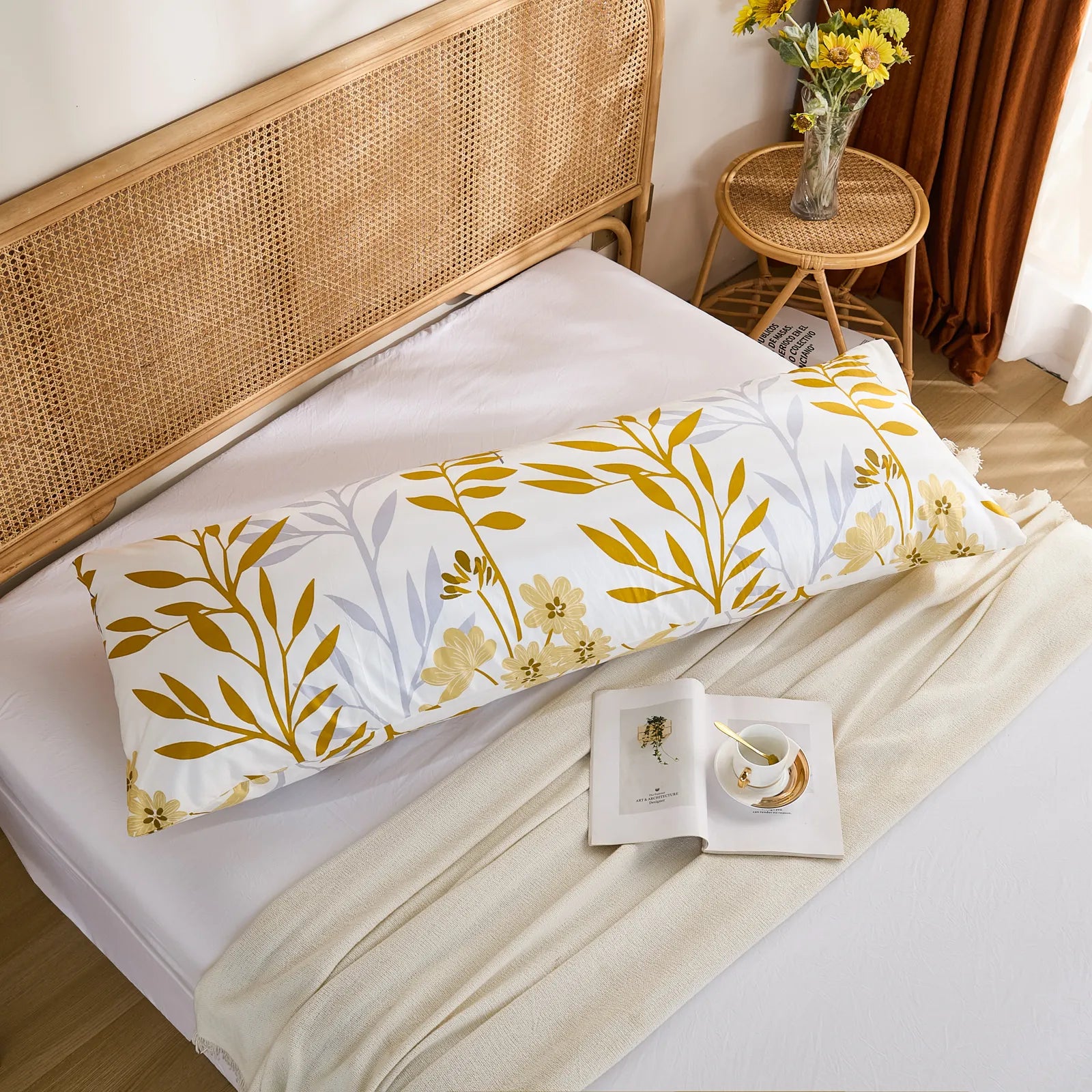A flat lay of a long white cotton body pillow, showcasing a bold yellow leaf and delicate floral pattern on a bed with a white blanket.