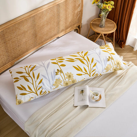 A flat lay of a long white cotton body pillow, showcasing a bold yellow leaf and delicate floral pattern on a bed with a white blanket.