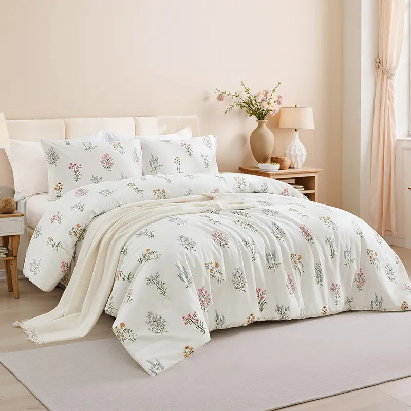 Full view of a bed with a white floral comforter and pillows, a wood headboard, and bedside table with a lamp