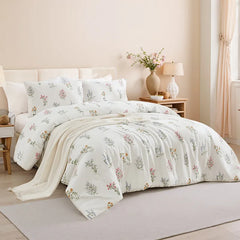 Full view of a bed with a white floral comforter and pillows, a wood headboard, and bedside table with a lamp