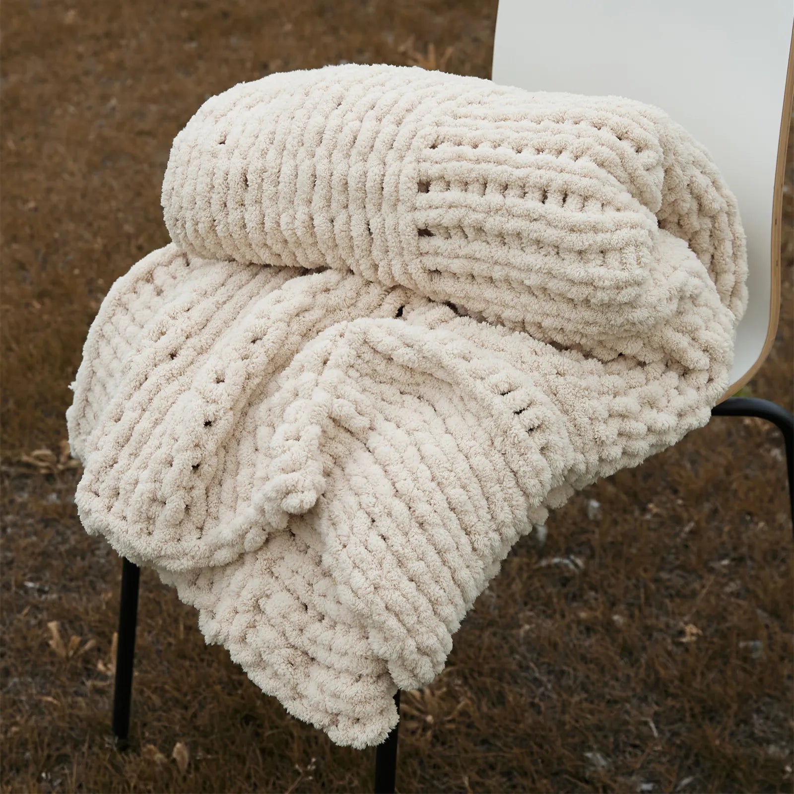 A close-up of a neatly folded beige chunky knit blanket on a chair, showcasing its thick, woven texture in an outdoor setting.