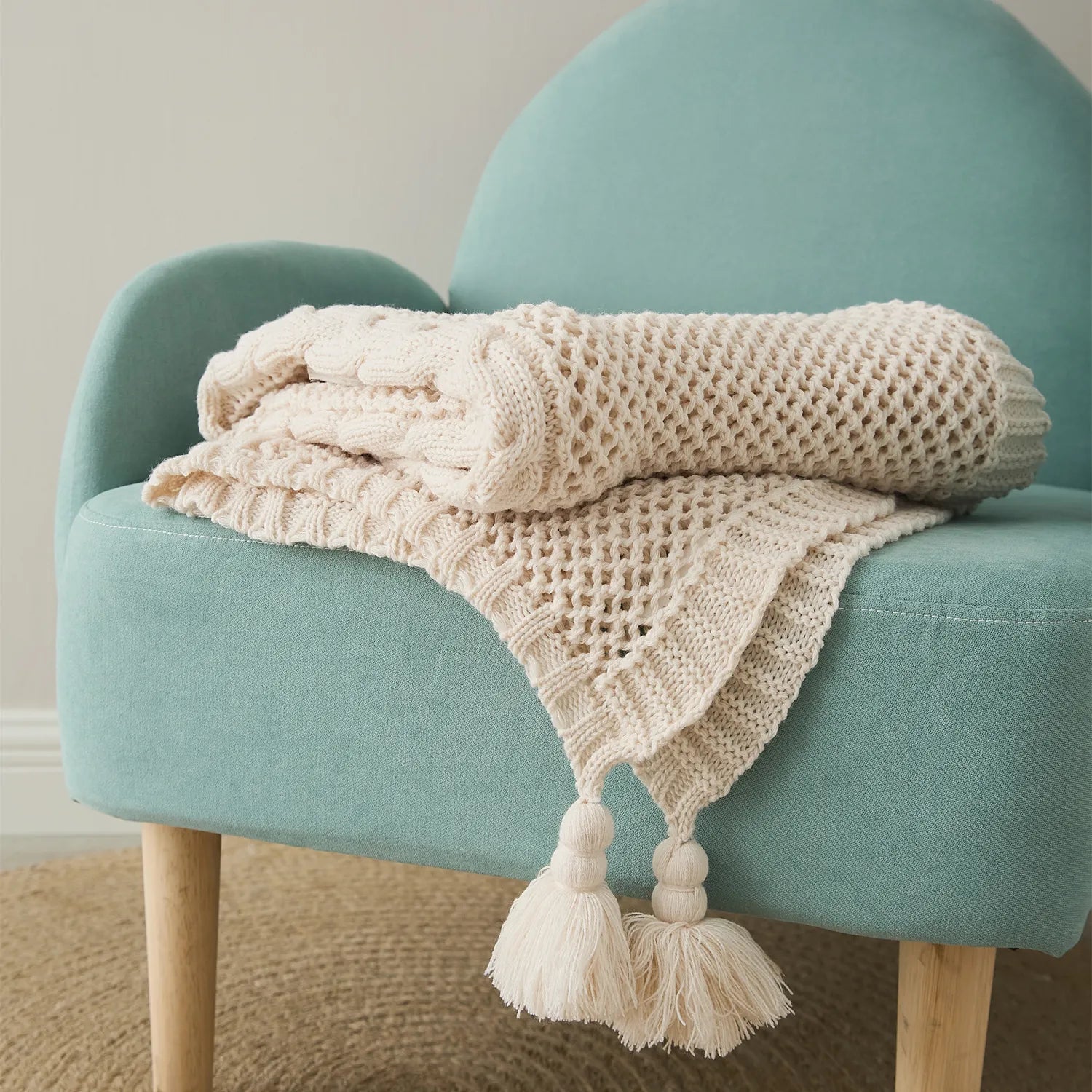 Folded cream-white knit throw blanket with tassels neatly placed on a modern teal armchair.