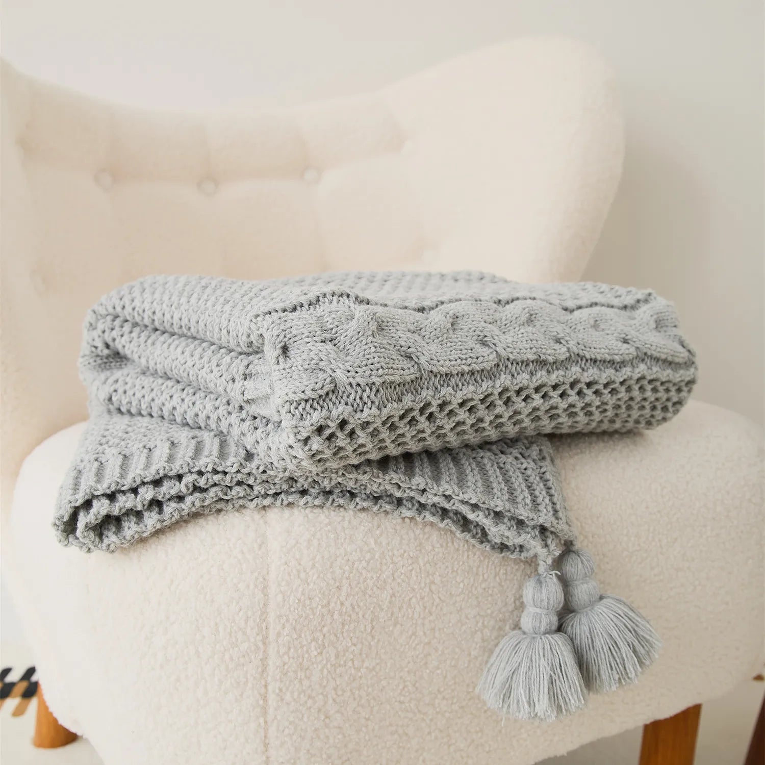 Folded light-grey cable-knit throw blanket with tassels neatly placed on a cream armchair.
