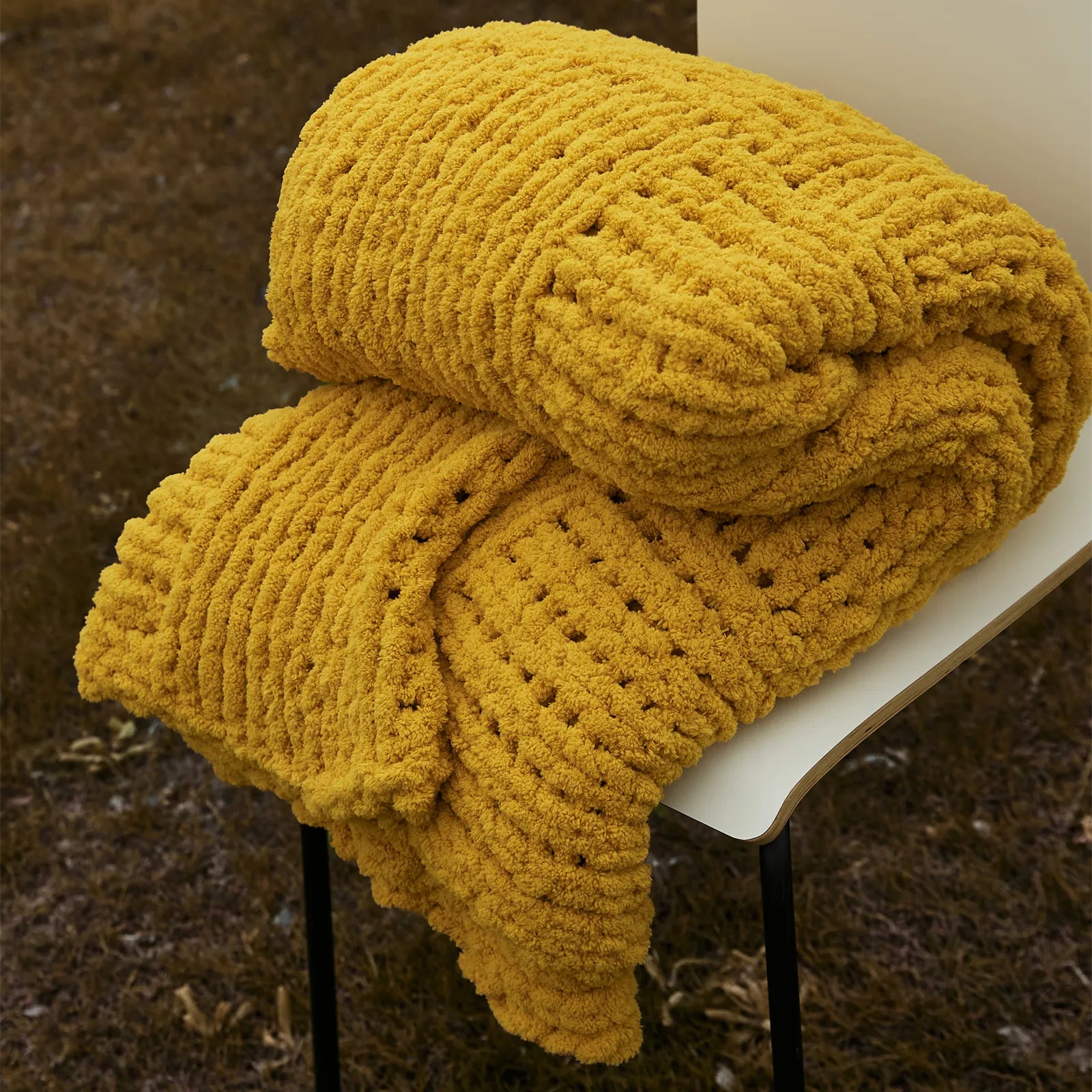 A folded mustard yellow chunky knit blanket is placed on a chair outdoors, showing its soft, plush texture.