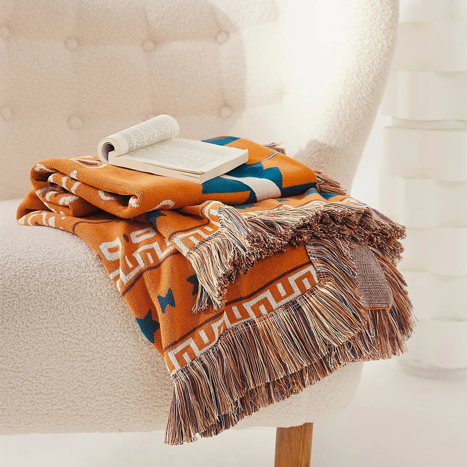 Folded orange bohemian jacquard cotton throw blanket with fringes and open book on a white armchair.
