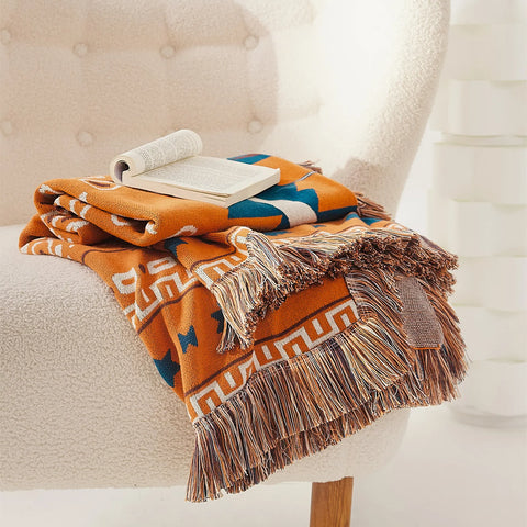 Folded orange bohemian jacquard cotton throw blanket with fringes and open book on a white armchair.