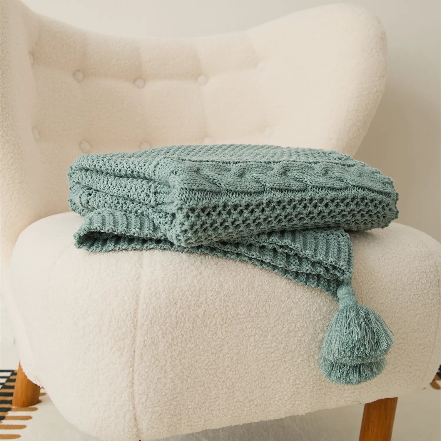 Folded sage-green cable-knit throw blanket with tassels on a cream armchair.