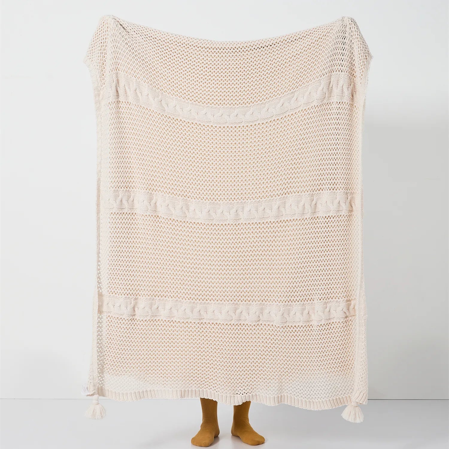 Full view of the large cream-white open-knit throw with horizontal cable stripes and tassels held up.