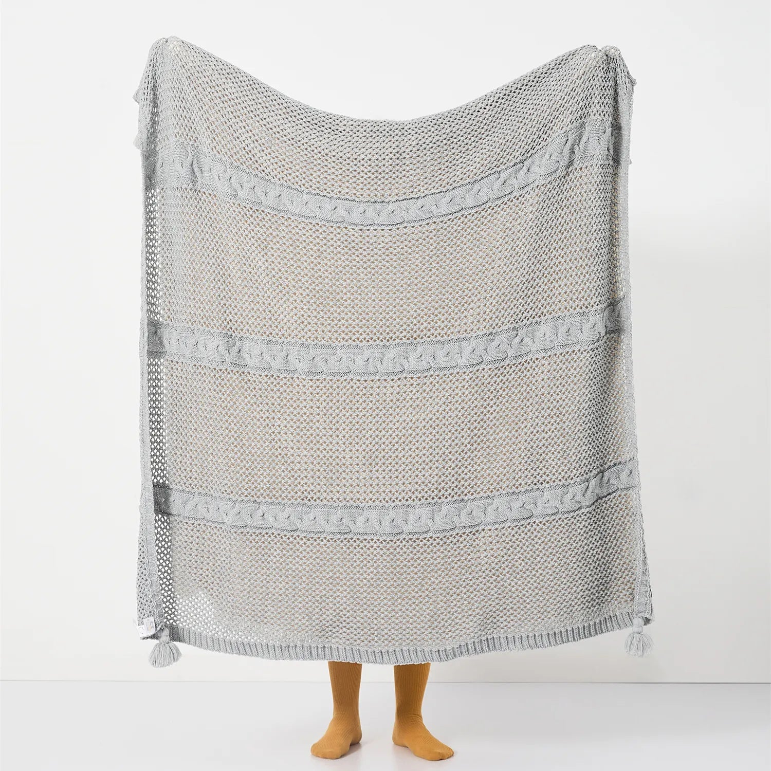 Full view of the large light-grey open-knit throw with horizontal cable stripes and tassels held up.
