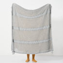 Full view of the large light-grey open-knit throw with horizontal cable stripes and tassels held up.
